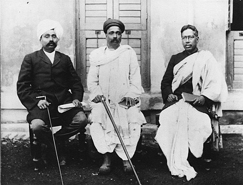 Black and white photograph of three seated men outside a building with louvred shutters: Lala Lajpat Rai in a light turban and dark coat, Bal Gangadhar Tilak in a turban and white dhoti holding a stick, and Bipin Chandra Pal in spectacles with a shawl across his shoulder.